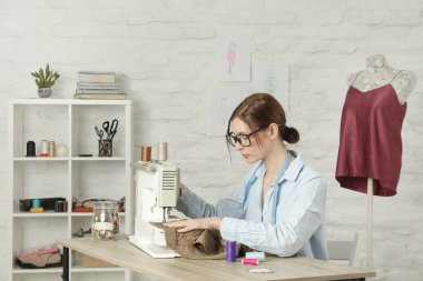 Küçük stüdyoda dikiş makinesinde dikiş diken bir kadın, moda atölyesi, yavaş moda, terzi işi, el yapımı kıyafetler.