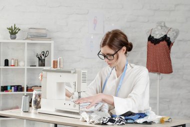Küçük stüdyoda dikiş makinesinde dikiş diken bir kadın, moda atölyesi, yavaş moda, terzi işi, el yapımı kıyafetler.