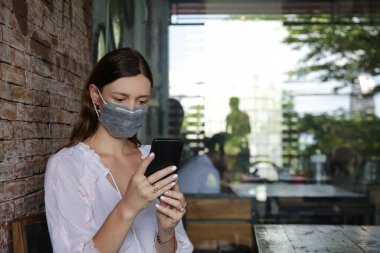 Bulaşıcı hastalıkları önlemek ve cep telefonundan haber okumak için yüz koruyucu maske takan genç beyaz kadın. Roman Coronavirüsü (2019-nCoV) veya Wuhan Coronavirus salgını kavramından korkuyor.