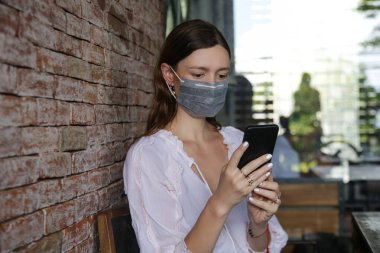 Bulaşıcı hastalıkları önlemek ve cep telefonundan haber okumak için yüz koruyucu maske takan genç beyaz kadın. Roman Coronavirüsü (2019-nCoV) veya Wuhan Coronavirus salgını kavramından korkuyor.
