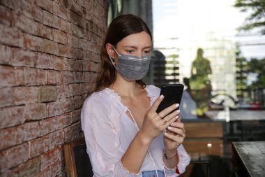 Bulaşıcı hastalıkları önlemek ve cep telefonundan haber okumak için yüz koruyucu maske takan genç beyaz kadın. Roman Coronavirüsü (2019-nCoV) veya Wuhan Coronavirus salgını kavramından korkuyor.