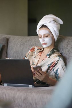 Laptopunun önünde yüzü güzellik maskesiyle oturan genç bir kadın. Evdeki sıcacık zaman. Kişisel zaman ve spa konsepti. Coronavirüs salgını ve kendini karantinaya alma kavramı.