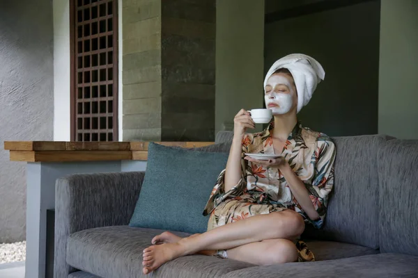 コーヒーを飲みながら顔の美容マスクで自宅に座っている若い女性 家での居心地の良い時間 セルフタイムとホームスパのコンセプト コロナウイルスの発生と自己隔離の概念 — ストック写真