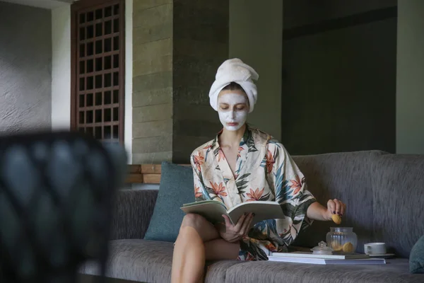 本を読みながら顔の美容マスクで家に座っている若い女性 家での居心地の良い時間 セルフタイムとホームスパのコンセプト コロナウイルスの発生と自己隔離の概念 — ストック写真