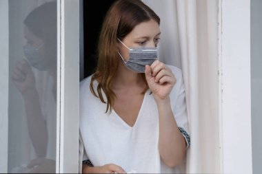 Ameliyat maskesiyle pencereden dışarı bakan genç bir kadın. Sıkıldım ve evde yapayalnızım. Coronavirüs salgını ve kendini karantinaya alma kavramı.