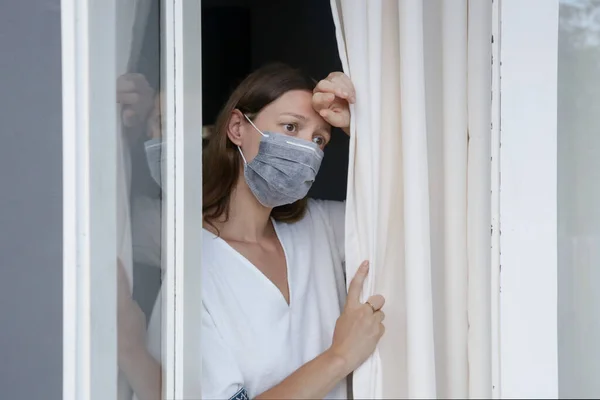 年轻女人带着外科一次性口罩透过窗户往外看 厌倦了一个人呆在家里头孢病毒的爆发和自我隔离的概念 — 图库照片