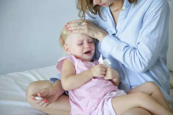 忧心忡忡的母亲 带着温度计坐在床上 抱着她那蹒跚学步的小女孩 母亲测量她生病孩子的体温 感冒和流感概念 — 图库照片