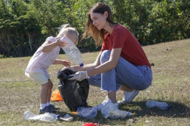 Genç bir kadın küçük kızıyla çayırdan plastik atık topluyor. Çöp toplamak, çocukları çevrenin korunmasına dahil etmek için yapılan eğitici bir faaliyettir..