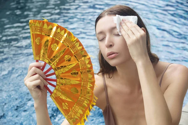 Woman fighting heat wave with a fan. Portrait of a young woman next to the water during a hot summer day.