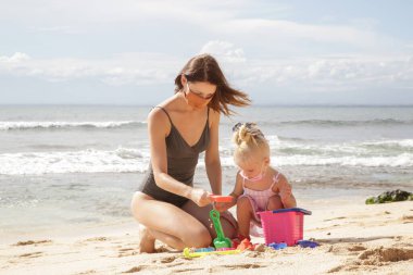 Coronavirus salgınından sonra yaz tatili. Sevimli küçük kız ve annesi kumsalda plaj oyuncaklarıyla oynarken koruyucu maske takıyor..
