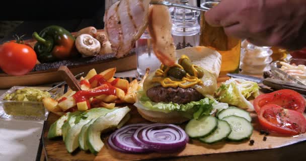 Añadir tocino crujiente sobre una hamburguesa con patatas fritas y cerveza — Vídeo de stock