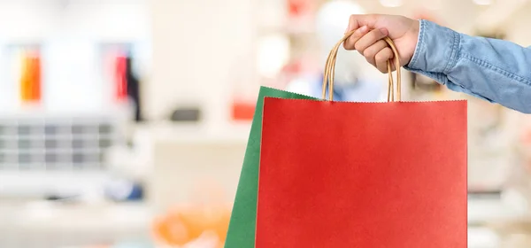 Ręka trzyma czerwona torba na zakupy na rozmycie tła sklepu, w baner — Zdjęcie stockowe