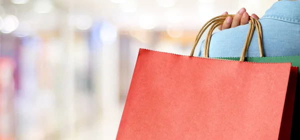 Ręka trzyma czerwona torba na zakupy na rozmycie tła sklepu, w baner — Zdjęcie stockowe