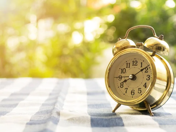 Vintage alarm clock on blue plaid tablecloth. The background is green from tree and light bokeh — Stock Photo, Image