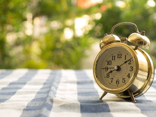 Vintage alarm clock on blue plaid tablecloth. The background is green from tree and light bokeh — Stock Photo, Image