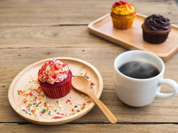 Kırmızı Top kek küresel ahşap tabağa koy. Yanında cupcake beyaz kahve kupa var. Arka planda sarı kek ve çikolatalı kek var. Hepsini tahta masada duruyor.