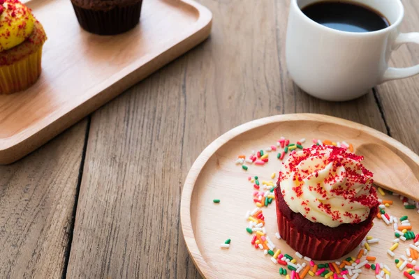 Cupcakes rouges mis sur une assiette en bois sphérique. À côté de cupcake ont tasse de café blanc. En arrière-plan ont cupcake jaune et cupcake au chocolat. Tout repose sur une table en bois . — Photo