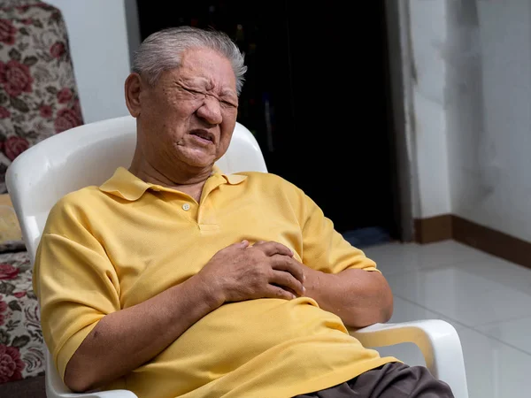 Homens asiáticos mais velhos sentados em uma cadeira na sala de estar com ataques cardíacos.Ambas as mãos do homem velho no peito por causa da respiração difícil, cara homem velho com dor causada por doença cardíaca . — Fotografia de Stock