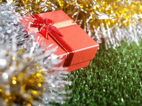 red gift box with red ribbon bow and golden seam place on silver and gold rainbow glowing decoration background on green grass. Christmas and New year concept.