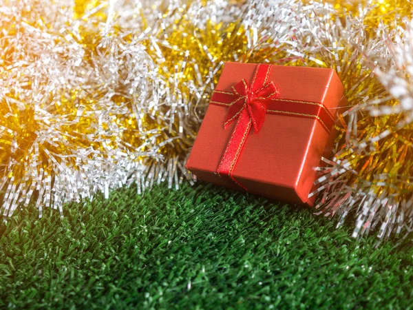 red gift box with red ribbon bow and golden seam place on silver and gold rainbow glowing decoration background on green grass. Christmas and New year concept.