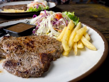 T-bone Steak tuz ve biber, altın kızarmış patates ve yeşil sebzeler ile kapatın. Beyaz tabak, çatal ve bıçak, restoranda ahşap masa