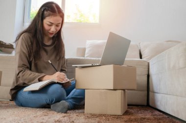 Genç girişimci, yerde oturuyor ve evde çalışıyor. Evet. Müşterilerin siparişlerini dizüstü bilgisayarla kontrol etmek ve müşteri adreslerini yazmak internet üzerinden sevkiyat yapmak. Küçük işletme sahibi kavramı.