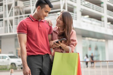 Alışveriş caddesinde renkli alışveriş torbalarını ellerinde tutarak yürüyen ve çek promosyonu için akıllı telefon kullanan mutlu genç çiftler. Satış ve Kara Cuma alışverişi kavramı