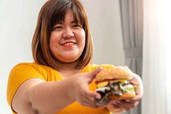 Hungry overweight woman smiling and holding hamburger and sitting in the bedroom, her very happy and enjoy to eat fast food. Concept of binge eating disorder (BED).