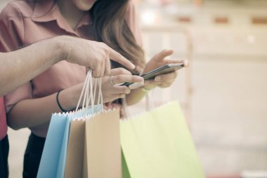 Alışveriş caddesinde renkli alışveriş torbalarını ellerinde tutarak yürüyen ve çek promosyonu için akıllı telefon kullanan mutlu genç çiftler. Satış ve Kara Cuma alışverişi kavramı