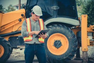 Mühendislik, kepçenin önünde duran beyaz bir kaskı takıyor ve planı inşaatla kontrol etmek için tablet kullanıyorlar.