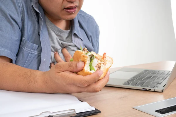 アジア系の男がオフィスの机の上でバーガーを食べて心臓発作 忙しいビジネスマンの概念は仕事とのバランスを取り 健康の世話をすることはできません唯一のジャンクフードを食べる — ストック写真