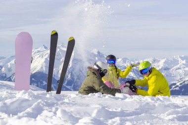 Üç mutlu arkadaş snowboardcular ve kayakçılar güneşli dağlarda öğle yemeği yiyorlar..