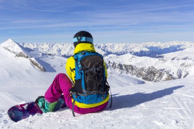 Snowboardcu sırtında bir yamaçta oturur ve dağların manzarasına bakar..