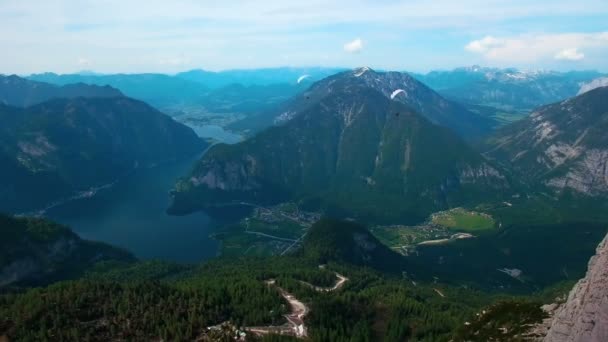 空中鸟瞰的飞行滑翔白色的降落伞从雪山在奥地利 在阿尔卑斯山的阳光明媚的时间 下面看到一个美丽的湖泊和乡村 — 图库视频影像