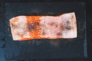 Yarım bir somon fileto ıslak bir taş tezgahının üzerine serpilmiş baharatlar ve tuz serpiştirilmiş.