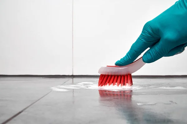 Mano masculina con cepillo rojo limpiando los azulejos del baño en el suelo . — Foto de Stock