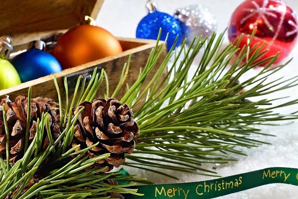 Weihnachtsgruß. Weihnachtskarte. ein Tannenzapfen auf einem Ast und eine Truhe voller Weihnachtskugeln auf schneebedecktem Hintergrund mit der Aufschrift Frohe Weihnachten. — Stockfoto