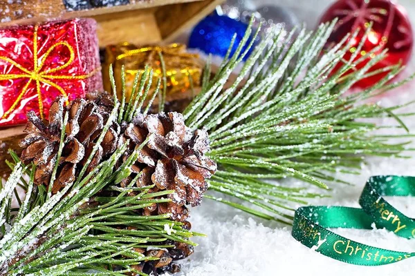 Ein Tannenzapfen auf einem Ast und eine Truhe voller Weihnachtskugeln auf schneebedecktem Hintergrund mit der Aufschrift "Frohe Weihnachten". Arrangement von Weihnachtsdekoration. — Stockfoto