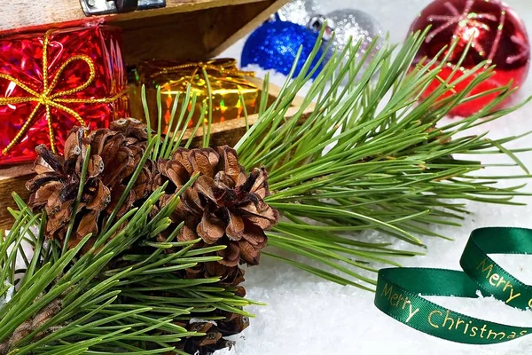 Ein Tannenzapfen auf einem Ast und eine Truhe voller Weihnachtsgeschenke auf schneebedecktem Hintergrund mit der Aufschrift "Frohe Weihnachten". Arrangement von Weihnachtsdekoration. — Stockfoto
