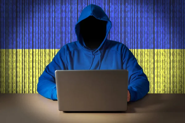 Hacker egy laptop előtt ülve, háttérben a digitális Ukrajna zászlaja. — Stock Fotó