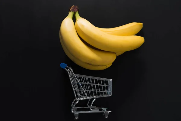 Bananas y carrito de compras sobre fondo negro . — Foto de Stock