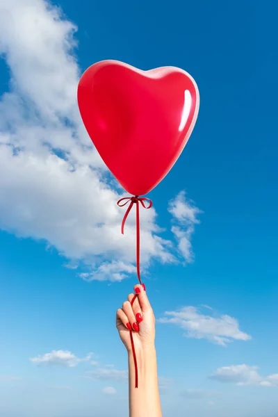 Womans hand with heart shaped balloon on background of sky.