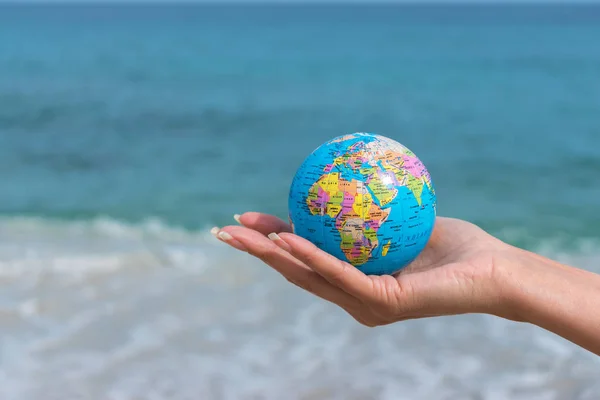 A mão humana está segurando um pequeno globo em frente ao mar. Viagens e questões globais conceito . — Fotografia de Stock