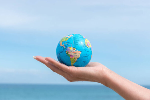 Human hand is holding small globe in front of the sea. 