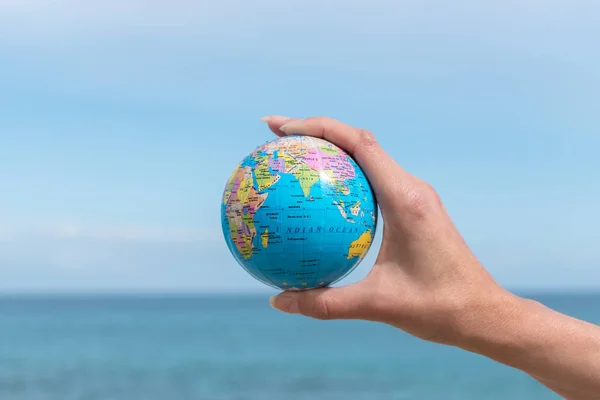 La main humaine tient un petit globe devant la mer. Concept Voyages et enjeux mondiaux . — Photo