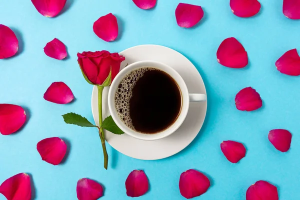 Creative composition of red rose flower and cup of coffee on blue background. Minimal spring concept.