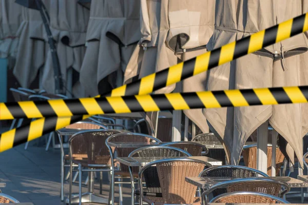 Barrier tape - quarantine, isolation concept, entry ban. Do not cross. Empty tables with umbrelas in restaurant on the street during sunrise.