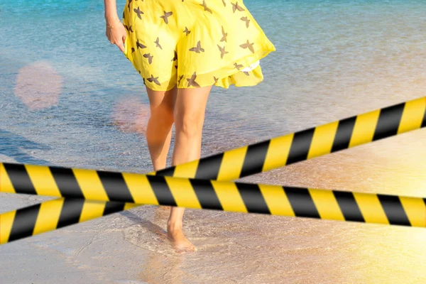 Barrier tape - quarantine, isolation concept, entry ban. Do not cross. A lonely girl is walking along island coastline and has reflection on wet sand.