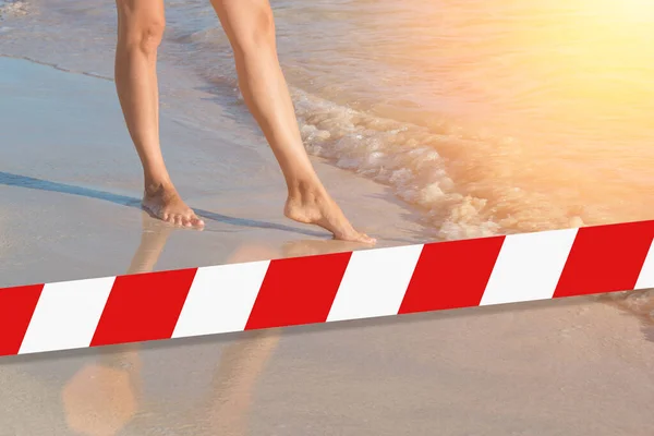 Barrier tape - quarantine, isolation concept, entry ban. Do not cross. A lonely girl is walking along island coastline and has reflection on wet sand.