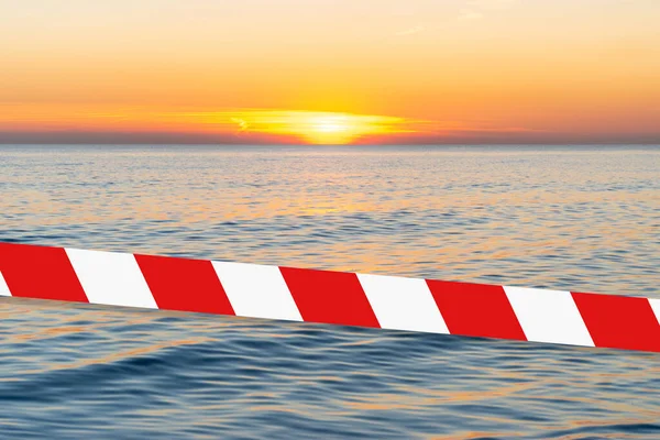 Barrier tape - quarantine, isolation concept, entry ban. Do not cross. Bright sunset with a yellow sun above the sea surface.